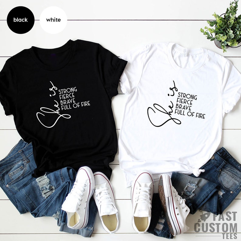 Pode incluir: Duas camisetas, uma preta e uma branca, com a palavra "She" escrita em letra cursiva e as palavras "Strong, Fierce, Brave, Full of Fire" empilhadas verticalmente abaixo.