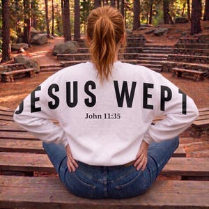 May include: White sweatshirt with the text "JESUS WEPT" in large black letters across the back, with "John 11:35" below. The sweatshirt is worn with blue jeans. The person is sitting on a wooden bench in a park.