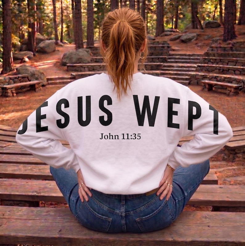 May include: White sweatshirt with the text "JESUS WEPT" in large black letters across the back, with "John 11:35" below. The sweatshirt is worn with blue jeans. The person is sitting on a wooden bench in a park.