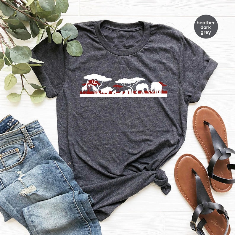 May include: Dark heather gray t-shirt featuring a white and red savanna graphic. The design includes silhouettes of giraffes, elephants, and trees. The text "heather dark grey" is in the upper right corner. The shirt is paired with blue jeans and sandals.