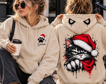 May include: A beige hooded sweatshirt featuring a black paw print with a red Santa hat and the name Emma on the front. The back displays a graphic of a black cat wearing a red Santa hat and scarf, with matching eye graphics on the hood.
