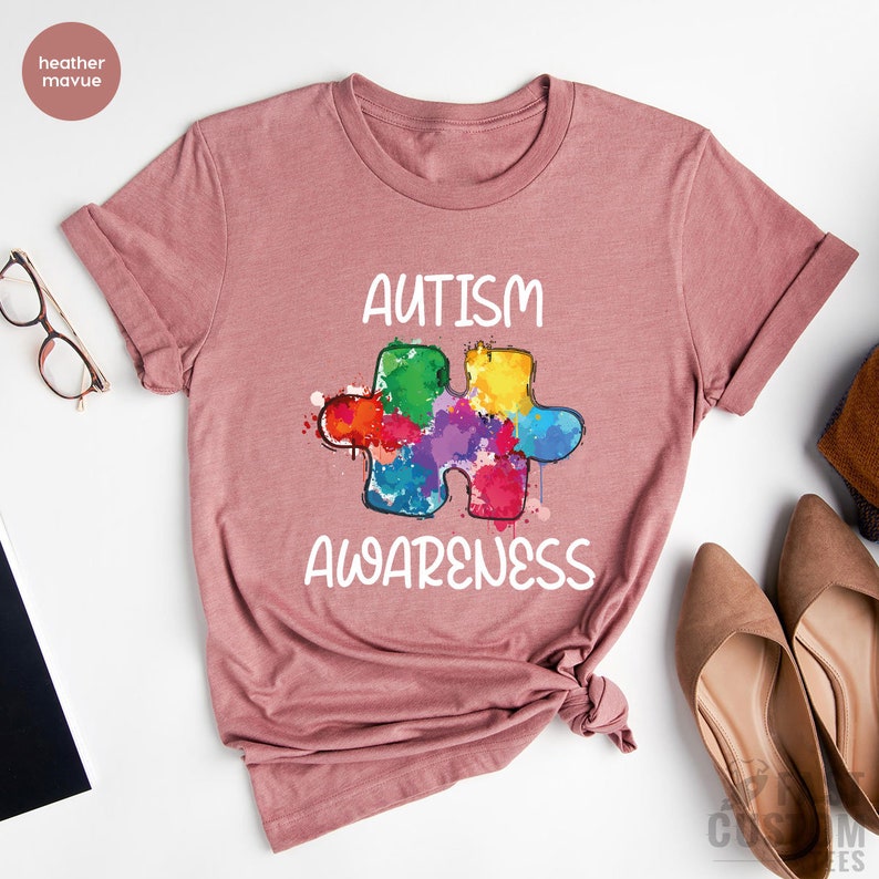 May include: A heather mauve t-shirt with the text "AUTISM AWARENESS" in white lettering. A colorful puzzle piece is centered on the shirt.