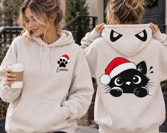 May include: A beige hooded sweatshirt featuring a black paw print with a red Santa hat and the name Emma on the front. The back displays a large graphic of a black cat wearing a red Santa hat, with cat ears printed on the hood.