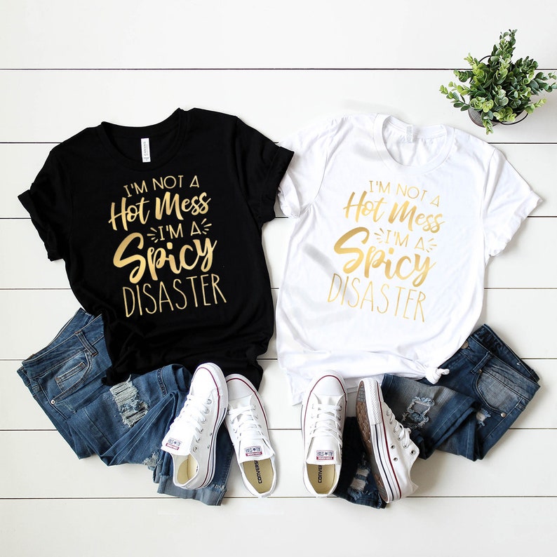 May include: Two t-shirts with the text "I'm not a hot mess I'm a spicy disaster" in gold lettering on a black and white shirt. The shirts are laid out on a white wooden surface with blue jeans and white sneakers.