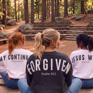May include: Three people wearing sweatshirts with inspirational messages. The center person's sweatshirt is dark gray with the word "FORGIVEN" and "Psalm 32:1". The other two sweatshirts are white, one says "PRAY CONTINU" and "1 Thessalonians 5:17", the other says "JESUS WEPT" and "John 11:35".