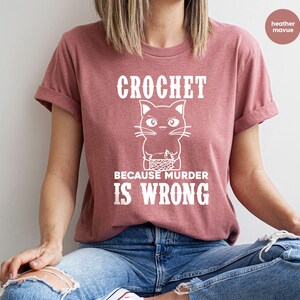 Op de afbeelding: Een heide-paarse t-shirt met een witte afbeelding van een breiende kat met de tekst "Crochet because murder is wrong."
