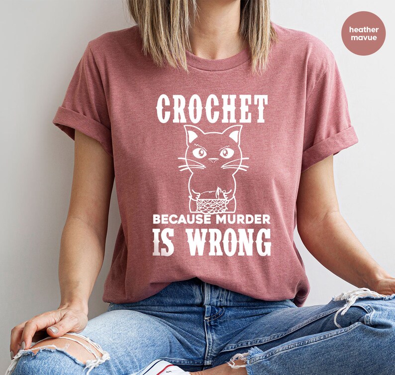 Op de afbeelding: Een heide-paarse t-shirt met een witte afbeelding van een breiende kat met de tekst "Crochet because murder is wrong."