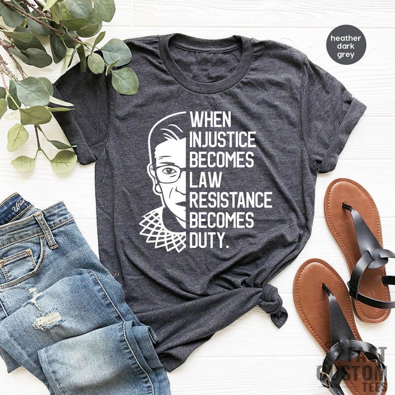 May include: A heather dark grey t-shirt with a white image of Ruth Bader Ginsburg and the text "When injustice becomes law resistance becomes duty."