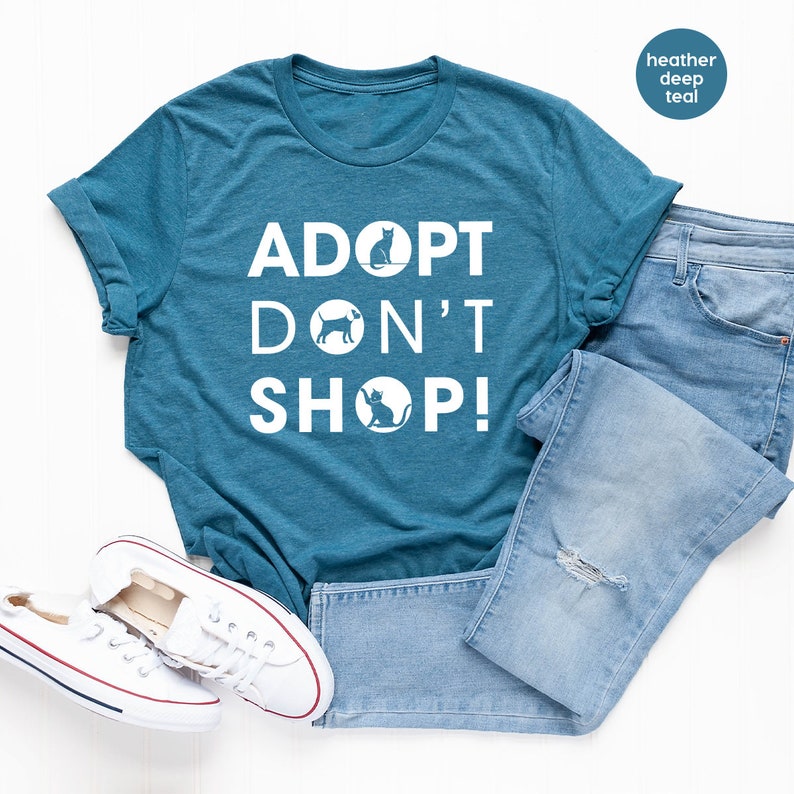 May include: A teal blue t-shirt with the text "ADOPT DON'T SHOP!" in white. The text is surrounded by white silhouettes of a cat and dog. The shirt is laid flat on a white surface with a pair of white sneakers and blue jeans.