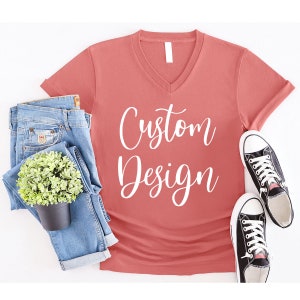 May include: A coral colored v-neck t-shirt with the text "Custom Design" in white lettering. The shirt is laying flat on a white surface with a pair of blue jeans and a potted plant.