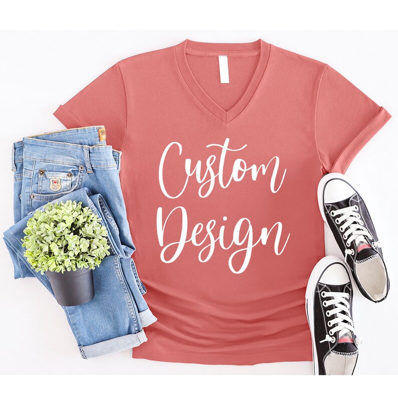 May include: A coral colored v-neck t-shirt with the text "Custom Design" in white lettering. The shirt is laying flat on a white surface with a pair of blue jeans and a potted plant.