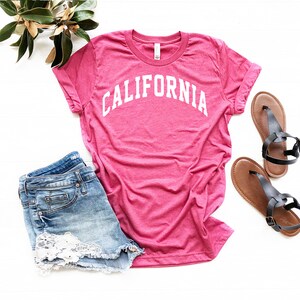 May include: A pink t-shirt with the word "CALIFORNIA" printed in white on the front. The shirt is paired with denim shorts and brown sandals.