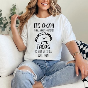 May include: White t-shirt with a black graphic that says "It's okay to fall apart sometimes tacos do and we still love them". The graphic features a cartoon taco with a smiling face.