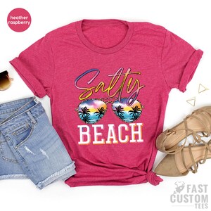 May include: A heather raspberry t-shirt with the text "Salty Beach" in a colorful, stylized font. The text is overlaid with a pair of sunglasses with a sunset and palm tree design.