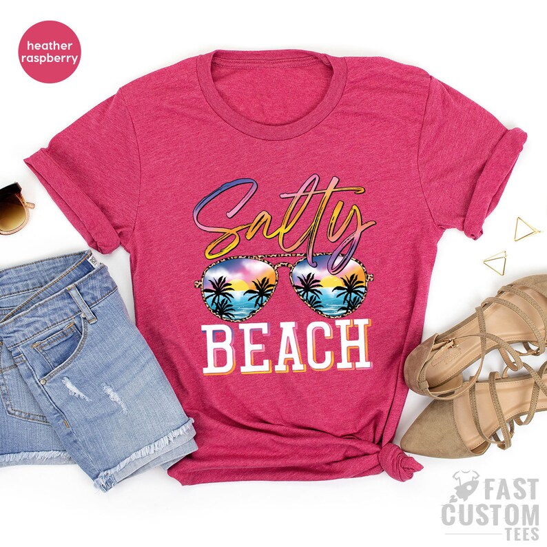 May include: A heather raspberry t-shirt with the text "Salty Beach" in a colorful, stylized font. The text is overlaid with a pair of sunglasses with a sunset and palm tree design.