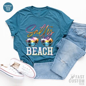 May include: A teal blue t-shirt with the text "Salty Beach" in a colorful, glittery font. The text is overlaid with a pair of sunglasses with a sunset and palm trees reflected in the lenses.