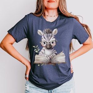 May include: A dark blue heathered t-shirt featuring a watercolour-style illustration of a young zebra wearing round glasses and reading an open book. The zebra has black and white stripes, a small tuft of hair on its head, and is surrounded by green leaves.
