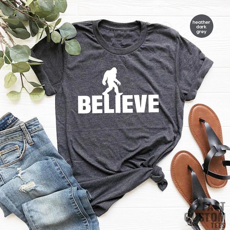 May include: A heather dark grey t-shirt with a white silhouette of Bigfoot walking and the word "BELIEVE" printed below it.