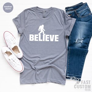 May include: A heather storm gray t-shirt with a white silhouette of Bigfoot walking and the word "BELIEVE" printed below it.