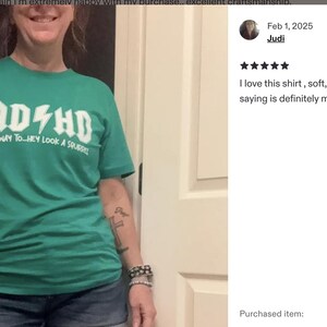 Op de afbeelding: Groen t-shirt met de witte tekst "ADHD Highway to Hey Look a Squirrel". Het t-shirt heeft een ronde hals en wordt door een persoon gedragen.