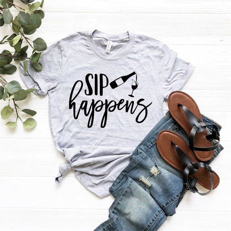 May include: A gray t-shirt with the text "SIP happens" in black lettering. A black silhouette of a wine bottle pouring into a wine glass is above the text.