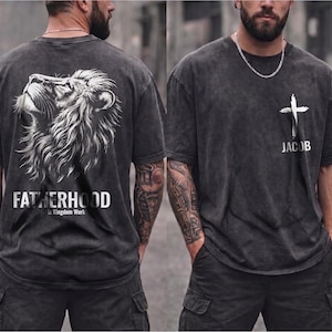 May include: Dark gray t-shirts with a distressed look. One shirt features a white lion graphic with the text "FATHERHOOD Is Kingdom Work." The other shirt has a white cross graphic and the name "JACOB."