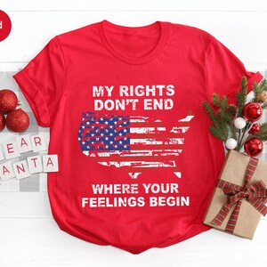 May include: A red t-shirt featuring the text "MY RIGHTS DON'T END WHERE YOUR FEELINGS BEGIN" and an American flag design in the shape of the United States. The shirt is shown with festive Christmas decorations.