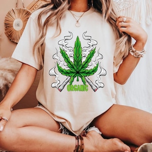 May include: Beige t-shirt featuring a green cannabis leaf design with two lit joints and the word "ORGANIC" in green. The graphic is centered on the shirt. The shirt is short-sleeved and has a crew neck.