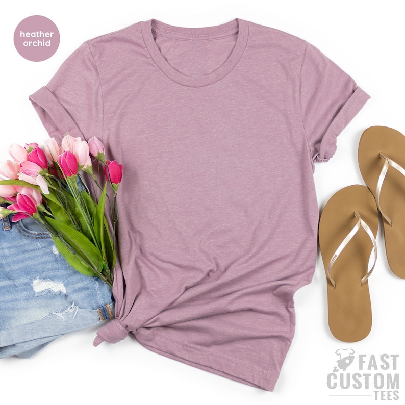 May include: A heather orchid colored t-shirt with rolled up sleeves. The t-shirt is laying on a white surface with a bouquet of pink tulips and a pair of brown flip flops.