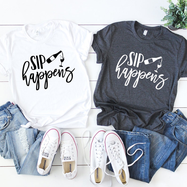 May include: Two t-shirts with the text "Sip Happens" and a graphic of a wine bottle pouring into a glass. The shirts are white and gray and are laid out on a white wooden surface with a pair of white sneakers and two pairs of blue jeans.