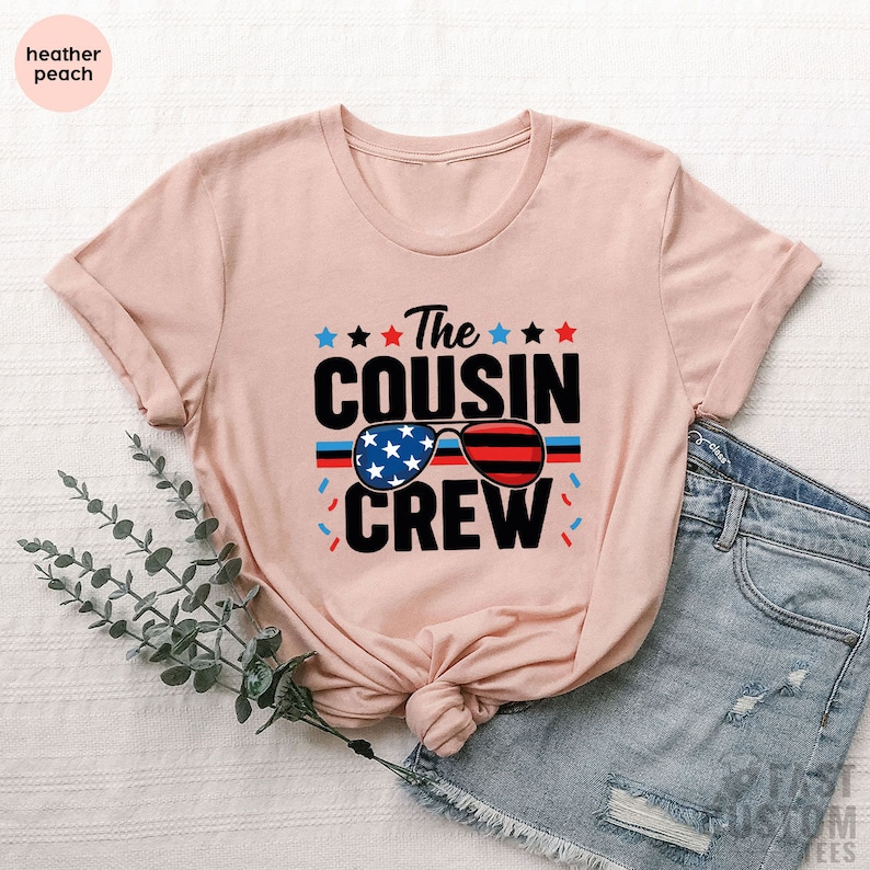 May include: A heather peach t-shirt with the text "The Cousin Crew" in black. The text is surrounded by stars and a graphic of sunglasses with an American flag design.