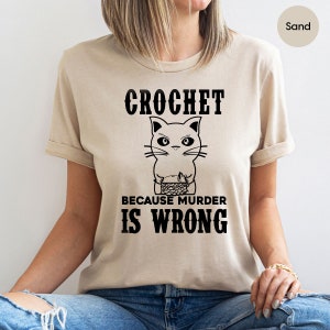 Op de afbeelding: Een beige t-shirt met een zwarte afbeelding van een kat die breit met de tekst "Crochet because murder is wrong".