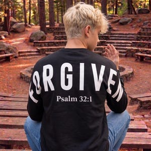 May include: Black long-sleeve shirt with the word "FORGIVEN" in large white letters across the back, along with "Psalm 32:1". The shirt is paired with blue jeans. The person is sitting on wooden steps in a forest setting.