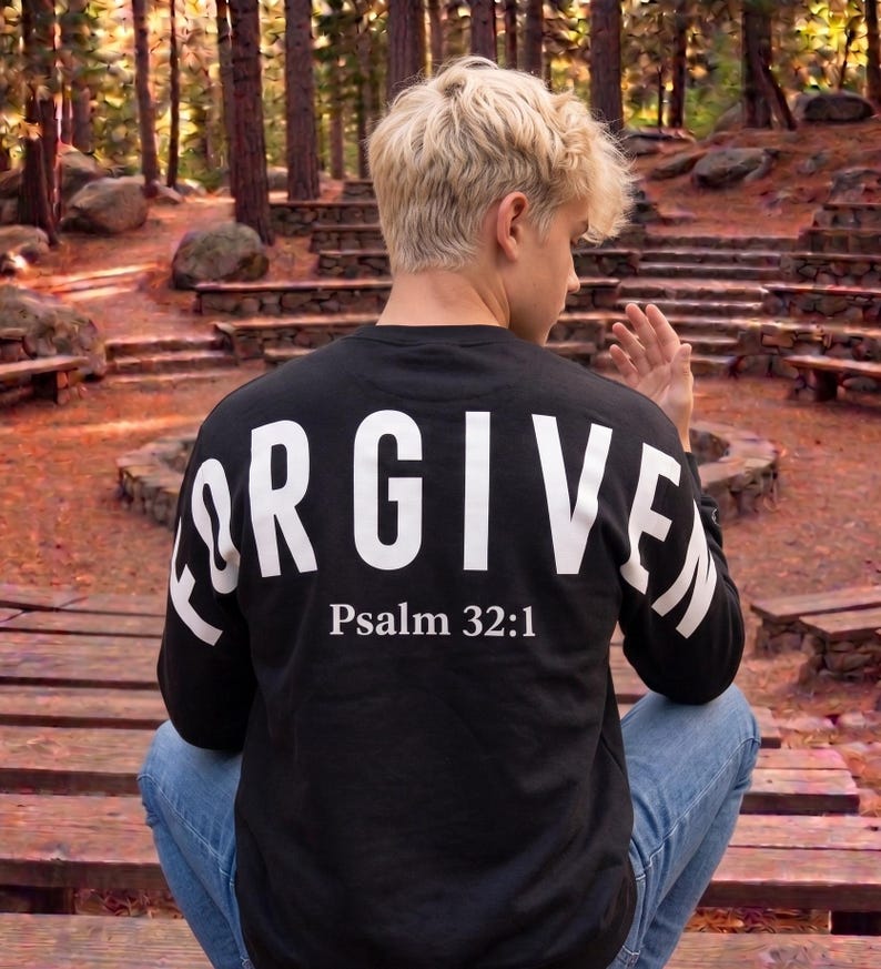 May include: Black long-sleeve shirt with the word "FORGIVEN" in large white letters across the back, along with "Psalm 32:1". The shirt is paired with blue jeans. The person is sitting on wooden steps in a forest setting.