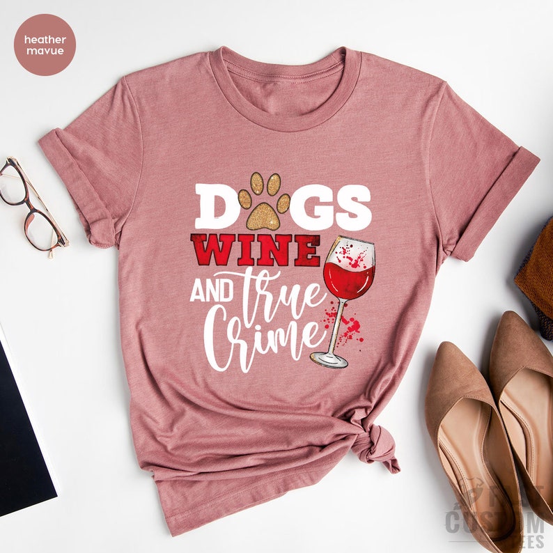 May include: A dusty rose t-shirt with the words "Dogs, Wine, and True Crime" printed on the front. The design includes a paw print, a wine glass, and a splash of red. The shirt is folded and has a knot at the bottom.