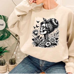 Puede incluir: Una sudadera de color arena con un estampado gr&aacute;fico de una mujer esqueleto con una bandana y flores. El gr&aacute;fico es en blanco y negro.