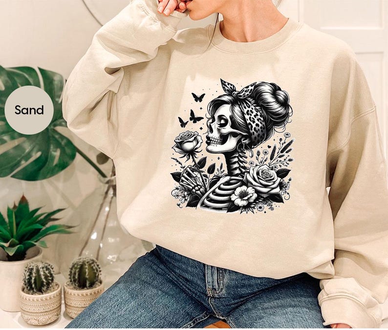 Puede incluir: Una sudadera de color arena con un estampado gr&aacute;fico de una mujer esqueleto con una bandana y flores. El gr&aacute;fico es en blanco y negro.