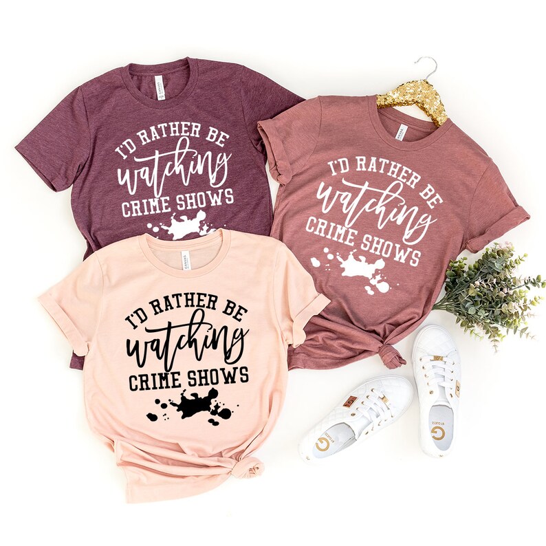 Pode incluir: Tr&ecirc;s t-shirts de manga curta nas cores bord&ocirc;, malva e p&ecirc;ssego. Cada t-shirt apresenta o texto branco ou preto "I'D RATHER BE watching CRIME SHOWS" com um gr&aacute;fico de salpicos de tinta por baixo. As t-shirts s&atilde;o exibidas com t&eacute;nis brancos.