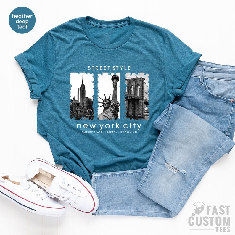 May include: A teal blue t-shirt with a graphic print of the Empire State Building, the Statue of Liberty, and the Brooklyn Bridge. The text "STREET STYLE" and "new york city" is printed on the shirt. The text "EMPIRE STATE - LIBERTY - BROOKLYN" is printed below the graphic.