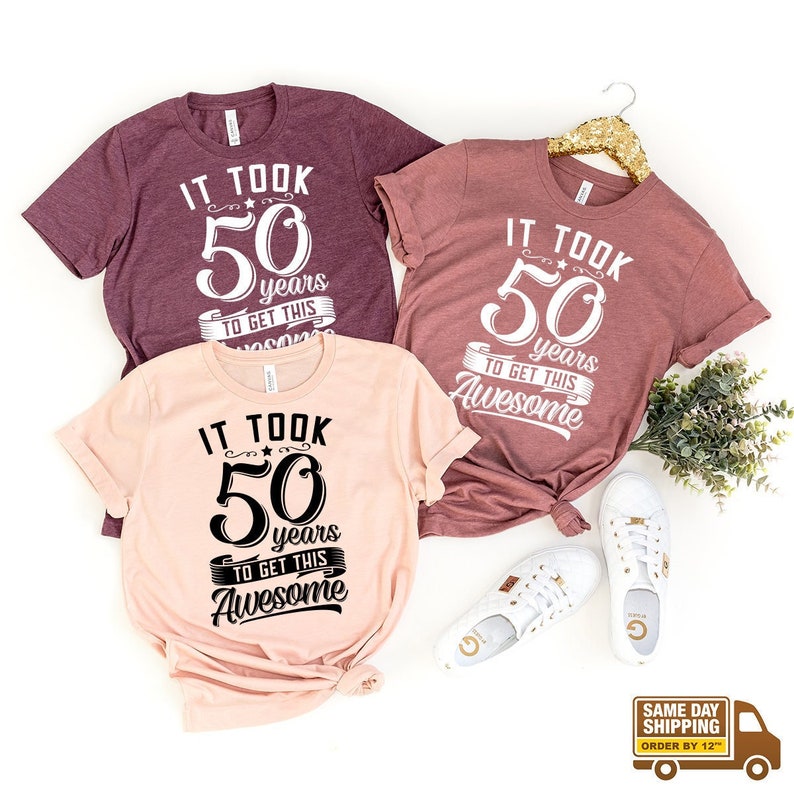 May include: Three t-shirts with the text "It took 50 years to get this Awesome" on them. The shirts are in different colors: maroon, pink, and peach.