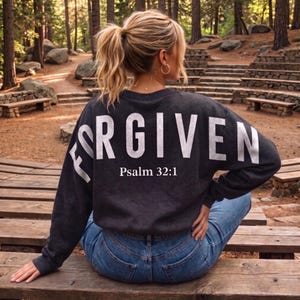 May include: Dark gray sweatshirt with the word "FORGIVEN" in large white letters across the back, with "Psalm 32:1" printed below. The sweatshirt is worn with blue jeans. The image is taken outdoors in a wooded area.