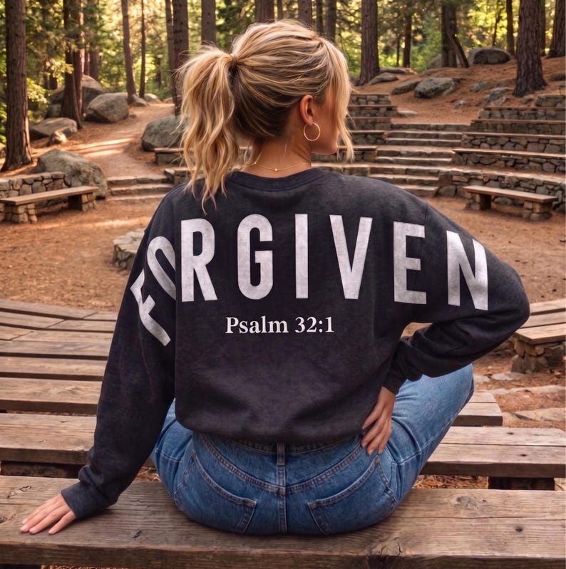May include: Dark gray sweatshirt with the word "FORGIVEN" in large white letters across the back, with "Psalm 32:1" printed below. The sweatshirt is worn with blue jeans. The image is taken outdoors in a wooded area.