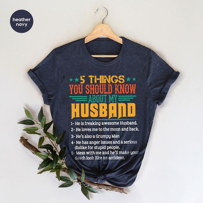 May include: A dark blue t-shirt with a white and orange graphic that says "5 Things You Should Know About My Husband". The graphic lists five things about the husband, including that he is "freaking awesome" and "loves me to the moon and back".