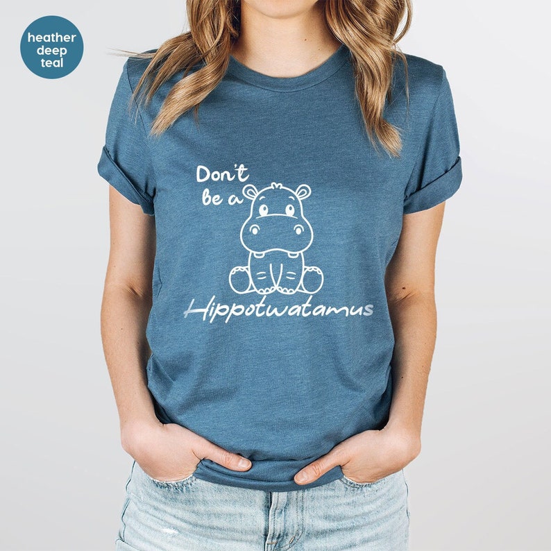 Pode incluir: Camiseta azul-petr&oacute;leo com o texto "Don't be a Hippotwatamus" e uma ilustra&ccedil;&atilde;o de hipop&oacute;tamo de desenho animado branco. A camisa tem mangas enroladas e gola redonda.