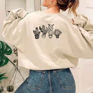 May include: Beige sweatshirt featuring a black graphic of four potted cacti and succulents. The design includes various cactus shapes and pot styles. The sweatshirt is a neutral color and has a relaxed fit, suitable for casual wear.