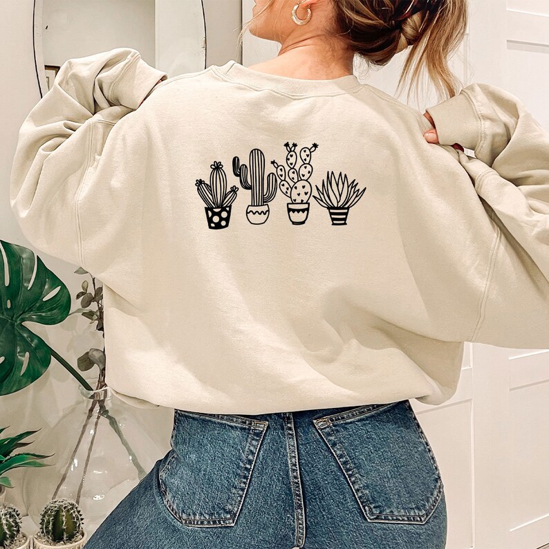 May include: Beige sweatshirt featuring a black graphic of four potted cacti and succulents. The design includes various cactus shapes and pot styles. The sweatshirt is a neutral color and has a relaxed fit, suitable for casual wear.