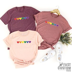 May include: Three t-shirts featuring a rainbow heart design. The shirts are in different colours: maroon, pink, and light pink.