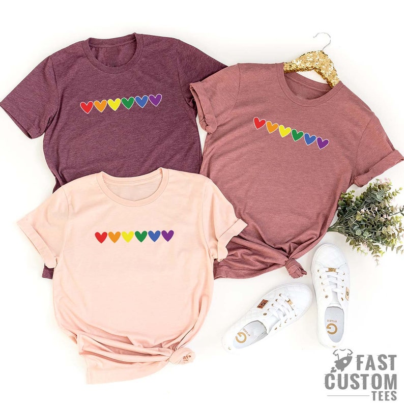 May include: Three t-shirts featuring a rainbow heart design. The shirts are in different colours: maroon, pink, and light pink.