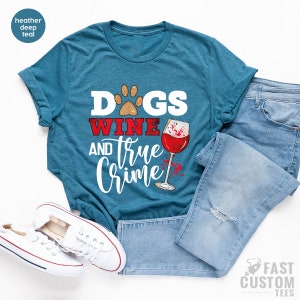 May include: Teal t-shirt with the text "Dogs Wine and True Crime" in white and red. The design includes a paw print, a wine glass with red wine, and a splatter effect. The shirt is paired with blue jeans and white sneakers.