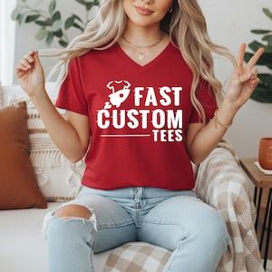 May include: A red V-neck t-shirt with the white text "FAST CUSTOM TEES" and a rocket ship graphic. The shirt is worn by a person wearing light blue jeans.